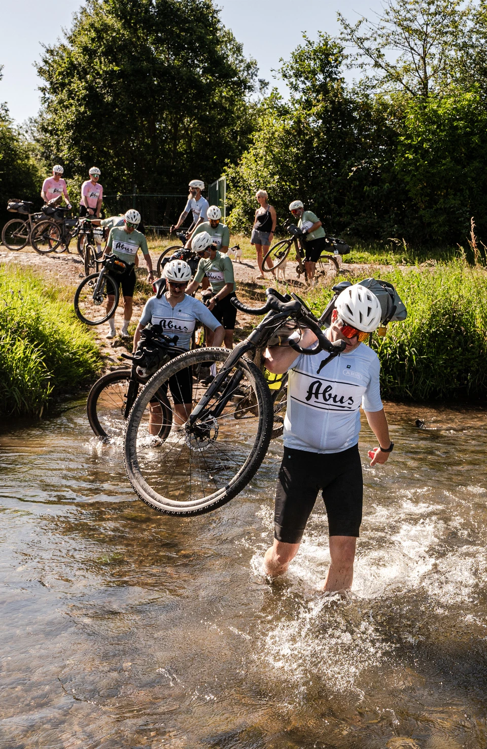 Un gruppo di ciclisti su strada trasporta le proprie biciclette attraverso un torrente durante un tour su ghiaia © ABUS