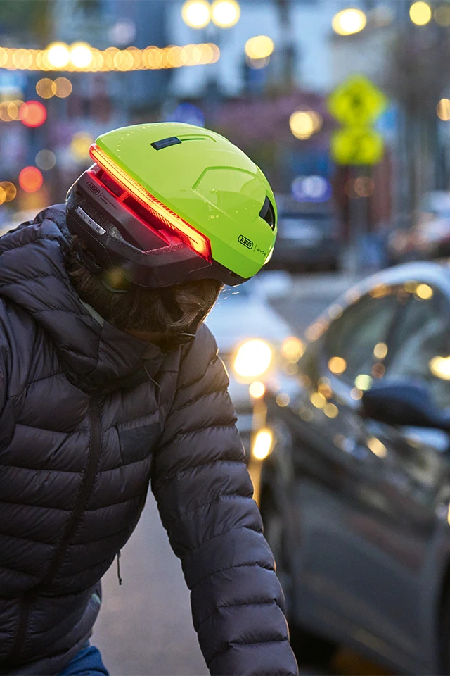 Mann fährt bei Dämmerung oder Abenddämmerung Fahrrad und trägt den gelben HYP-E Fahrradhelm mit integriertem Rücklicht. Der Helm ist mit einem roten LED-Rücklicht ausgestattet, das sichtbar leuchtet. Autos und Straßenlichter im Hintergrund leicht sichtbar © ABUS