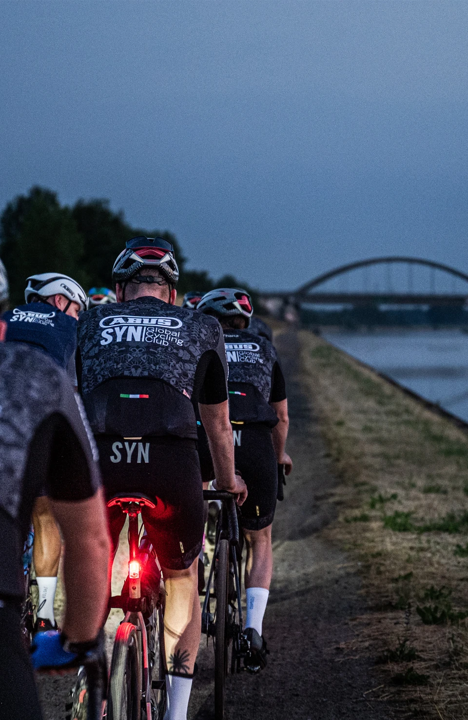 Ciclisti su strada pedalano in gruppo lungo la diga al tramonto © ABUS