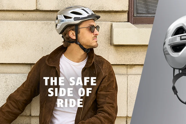 A man wearing an Urban-I 4.0 LR urban bike helmet in Signal Silver, seen from the side in front of a house wall © ABUS