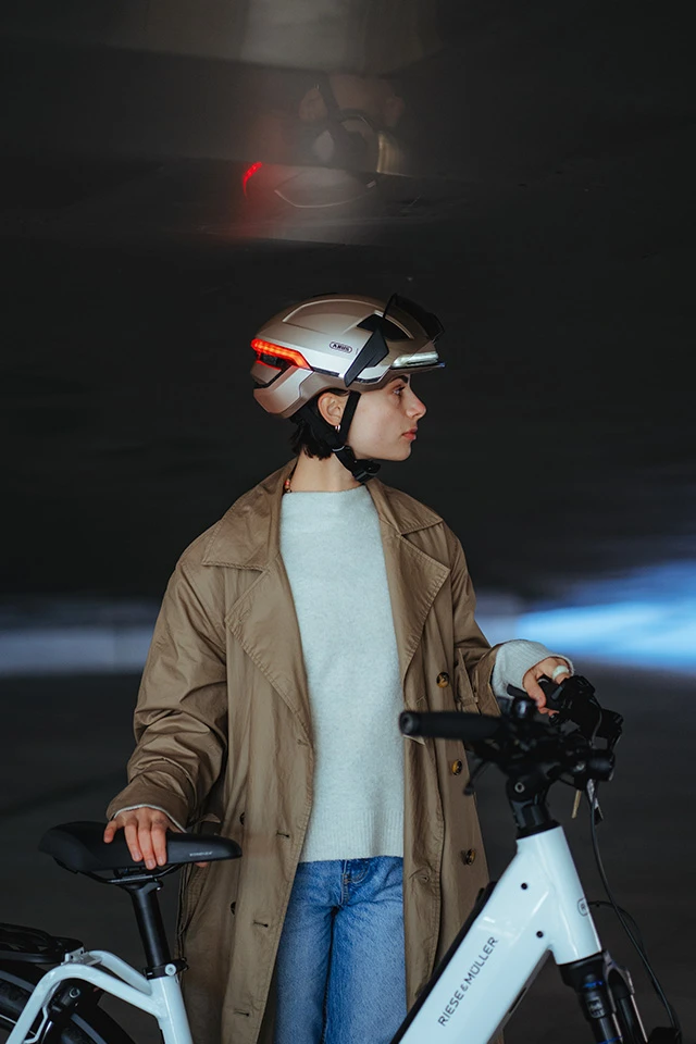 Eine Frau trägt den HYP-E Fahrradhelm in der Farbe gleam silver mit rotem Rücklicht und leuchtenden Frontlicht neben einem weißen E-Bike stehend © ABUS