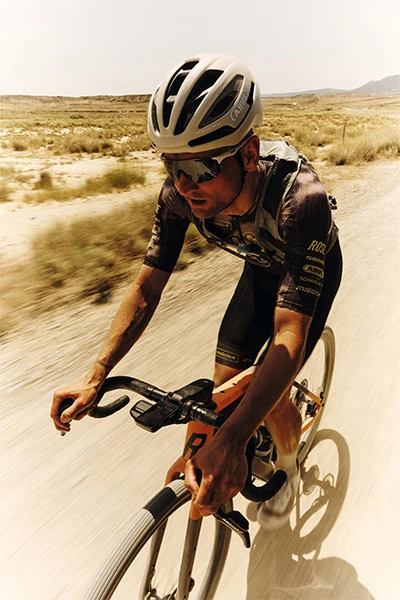Paul Voss porte un casque Taipan Gravel et roule à toute vitesse sur un chemin poussiéreux, entouré d'une végétation floue. © ABUS