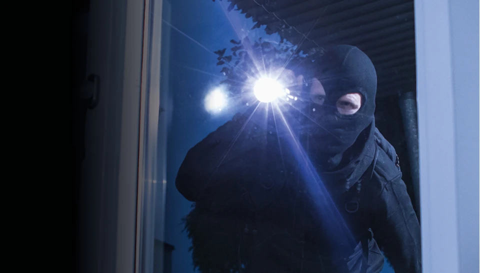 Großaufnahme eines Einbrechers mit Taschenlampe, der durch ein Fenster in eine Wohnung schaut.