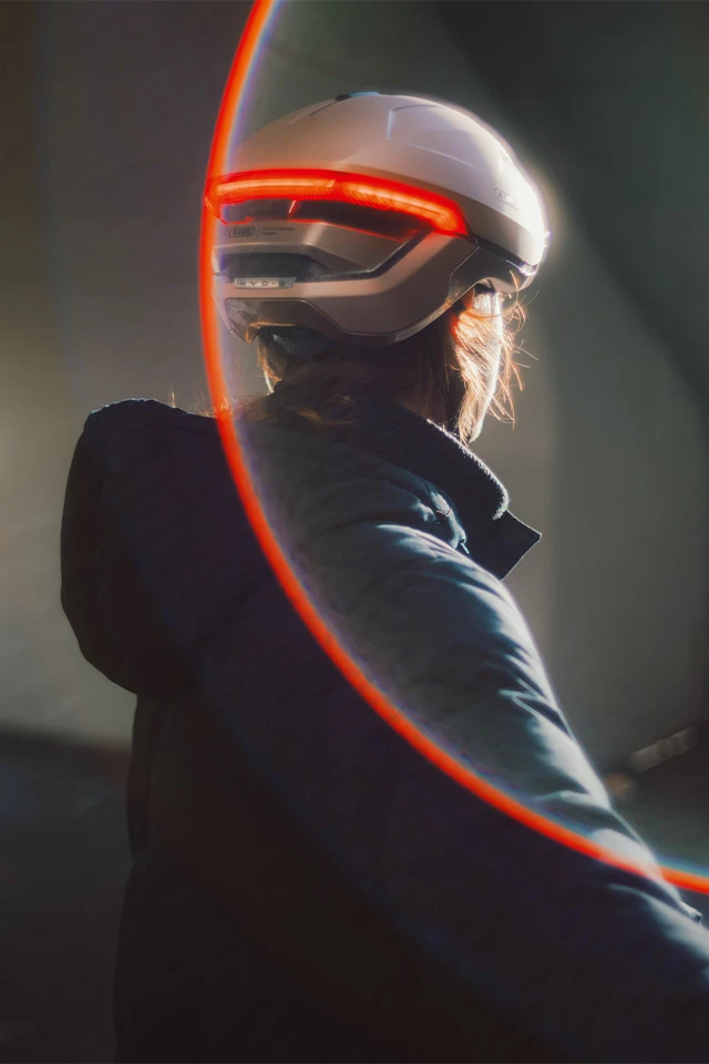 Rear view of the HYP-E bike helmet with red rear light, light flares, and a blurred background © ABUS