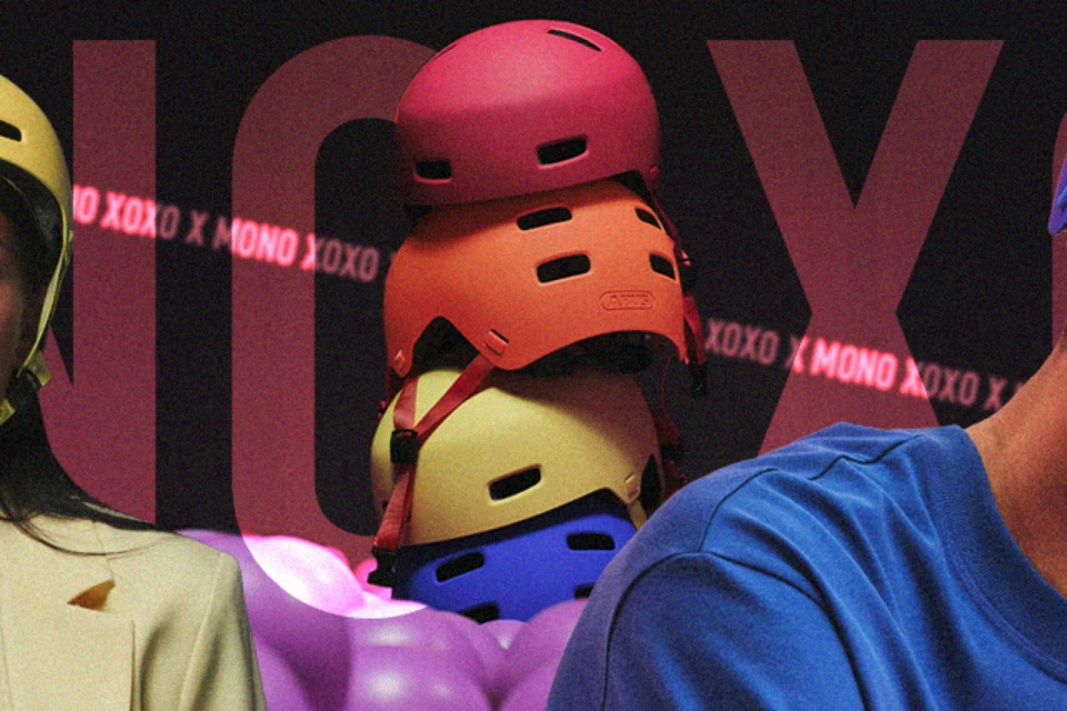 Gestapelte Fahrradhelme des Modells XOXO in den Farben Mono red, Mono orange, Mono yellow und Mono blue vor dunklem Hintergrund mit leuchtendem XOXO X MONO-Schriftzug © ABUS