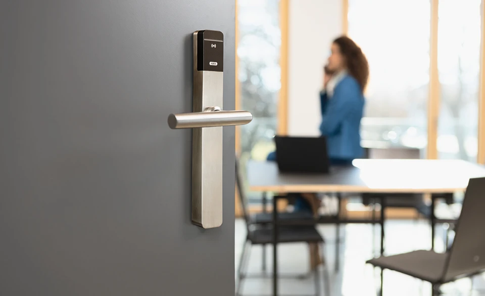 Halbgeöffnete Bürotür mit Zylinder, im Hintergrund eine Frau am Telefon