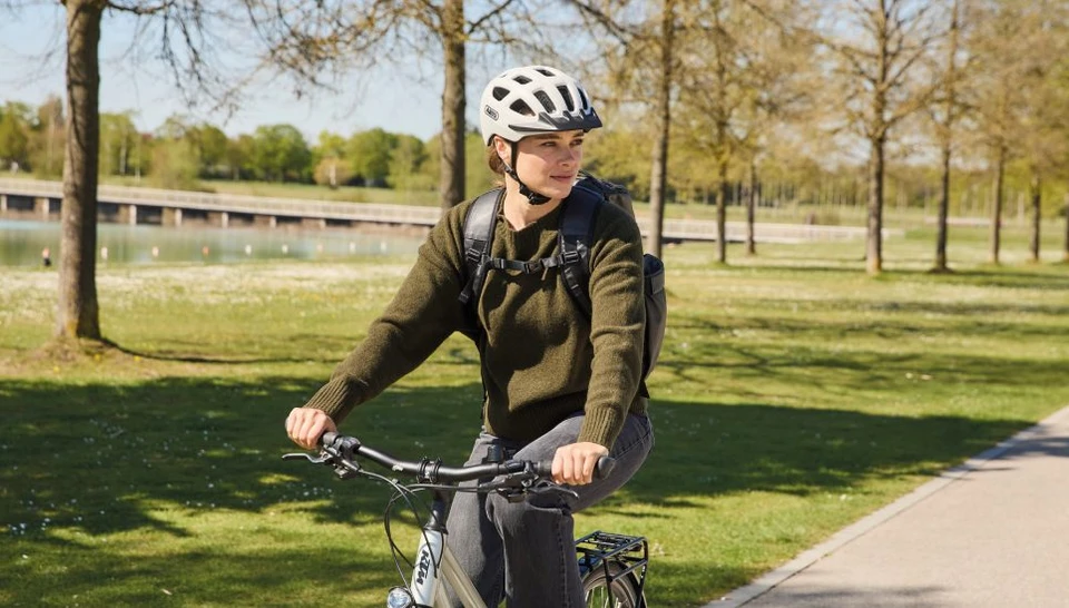 A woman is riding her bike along the lake and glances to the side. She is wearing the YOUN-I 3.0 ACE bike helmet in off-white © ABUS