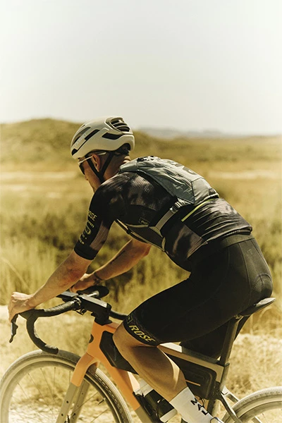 Paul Voss con il casco Taipan Gravel, visto da dietro, pedala su una strada sterrata chiara attraverso la campagna aperta © ABUS