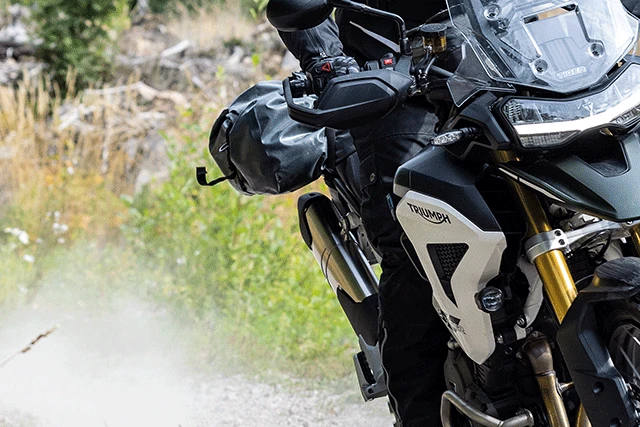 Un motard vêtu d'une combinaison noire conduit une moto enduro Triumph sur une piste de gravier hors route. ©ABUS