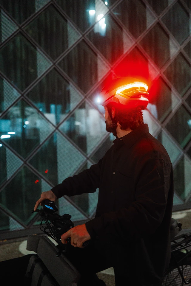Ein Mann trägt den HYP-E Fahrradhelm mit rotem Rücklicht und steht neben einem E-Bike. Der Helm leuchtet bei Nacht, während die Person vor einer modernen Glasfassade steht © ABUS