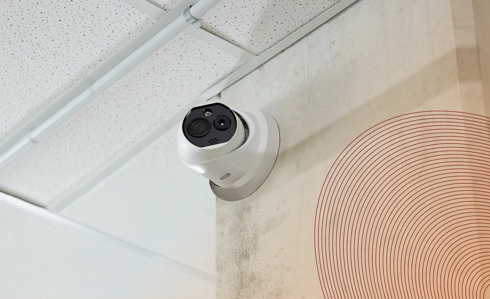 Weiße Kamera direkt unter der Decke an eine Wand montiert. Wand und Decke sind ebenfalls weiß.