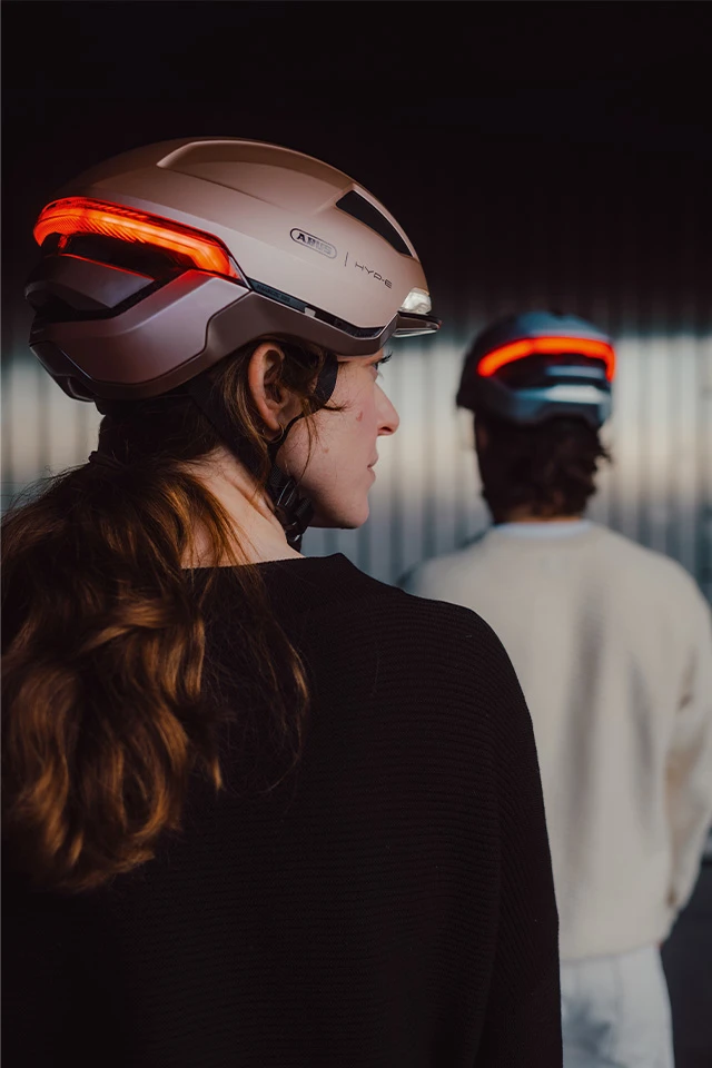 Deux personnes portant des casques de vélo HYP-E avec des feux arrière rouges, vues de dos — au premier plan en Champagne Gold, à l'arrière-plan en Midnight Blue © ABUS