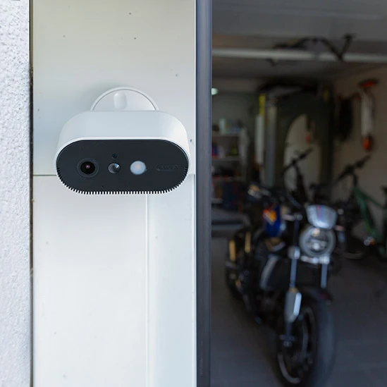 A white surveillance camera with integrated light is attached to a garage in which a motorbike is parked and observes the entrance area. ©ABUS