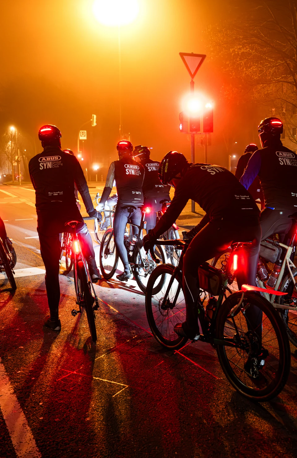 Ciclisti su strada che indossano caschi da bicicletta Taipan LED attendono a un incrocio di notte, con luci posteriori rosse che garantiscono la visibilità. © ABUS