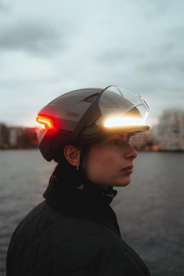 Seitliche Nahaufnahme einer Frau, die den HYP-E ACE Fahrradhelm mit integriertem Visier und rotem Rücklicht trägt. Der Helm leuchtet bei Dämmerung, und die Person blickt nach recht © ABUS