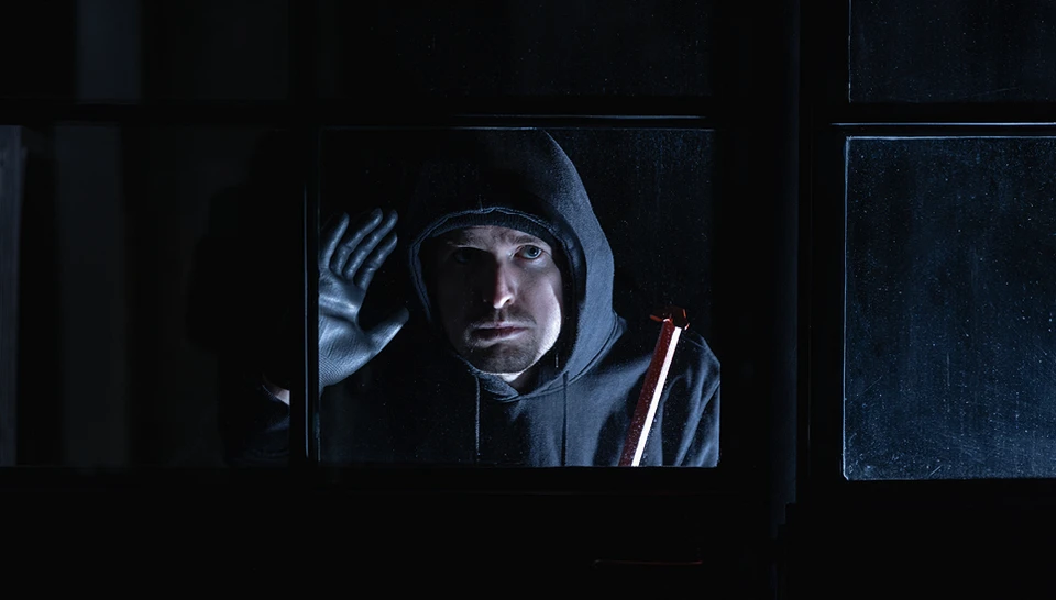 Ein schwarzgekleideter Einbrecher mit Brechstange späht durch ein Fenster ins Innere.