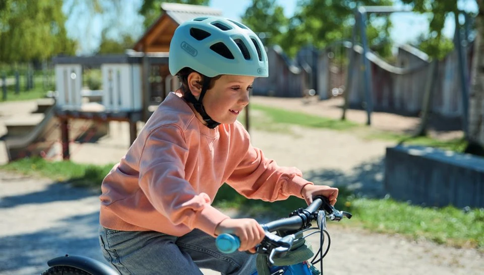 A young boy cycles past a playground; he is wearing the YOUN-I 3.0 powder blue cycle helmet © ABUS