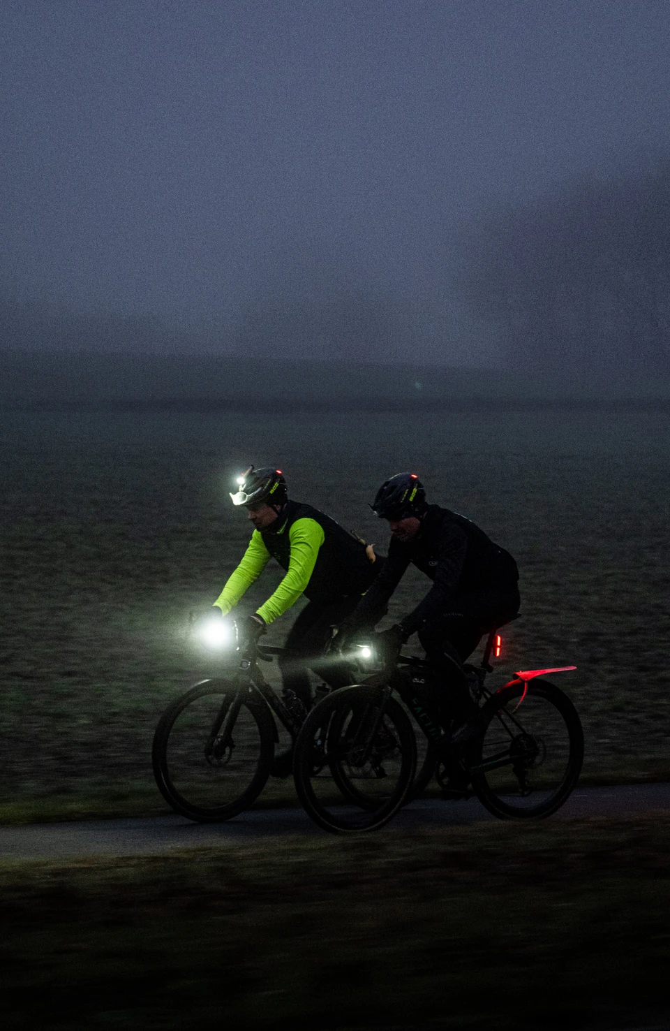 Two road cyclists wearing Taipan LED bicycle helmets ride along a country road in the dark with their lights switched on © ABUS