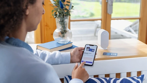 Frau h&auml;lt Smartphone mit ge&ouml;ffneter ABUS One App in der Hand w&auml;hrend vor ihr eine Keycard und eine Bridge One auf dem Sideboard stehen &copy; ABUS