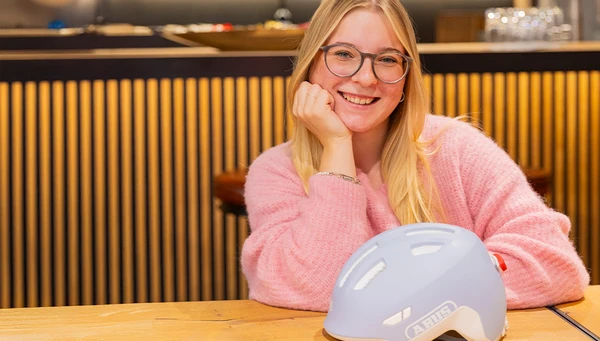 Produktmanagerin sitzt l&auml;chelnd an einem Tisch, der urbane Fahrradhelm HUD-Y in der Farbe lavender liegt vor ihr &copy; ABUS