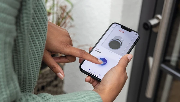 Vrouw houdt smartphone met ABUS One app in haar hand en opent de deur voor haar met de CYLOX One &copy; ABUS